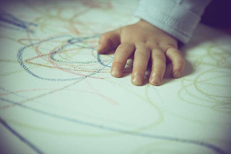 Dieses Bild zeigt die Hand eines Kleinkindes welche ein Blatt auf dem mit Wachsmalkreide gemalt ist berührt.